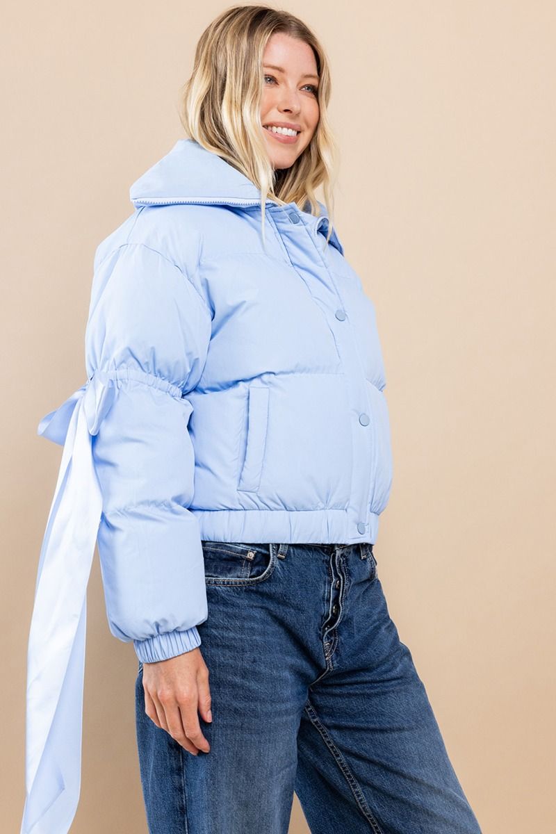 Collared, puffer jacket in baby blue with bow knot elbow string. Zipper and button closure. Featuring side pockets.  Side view.