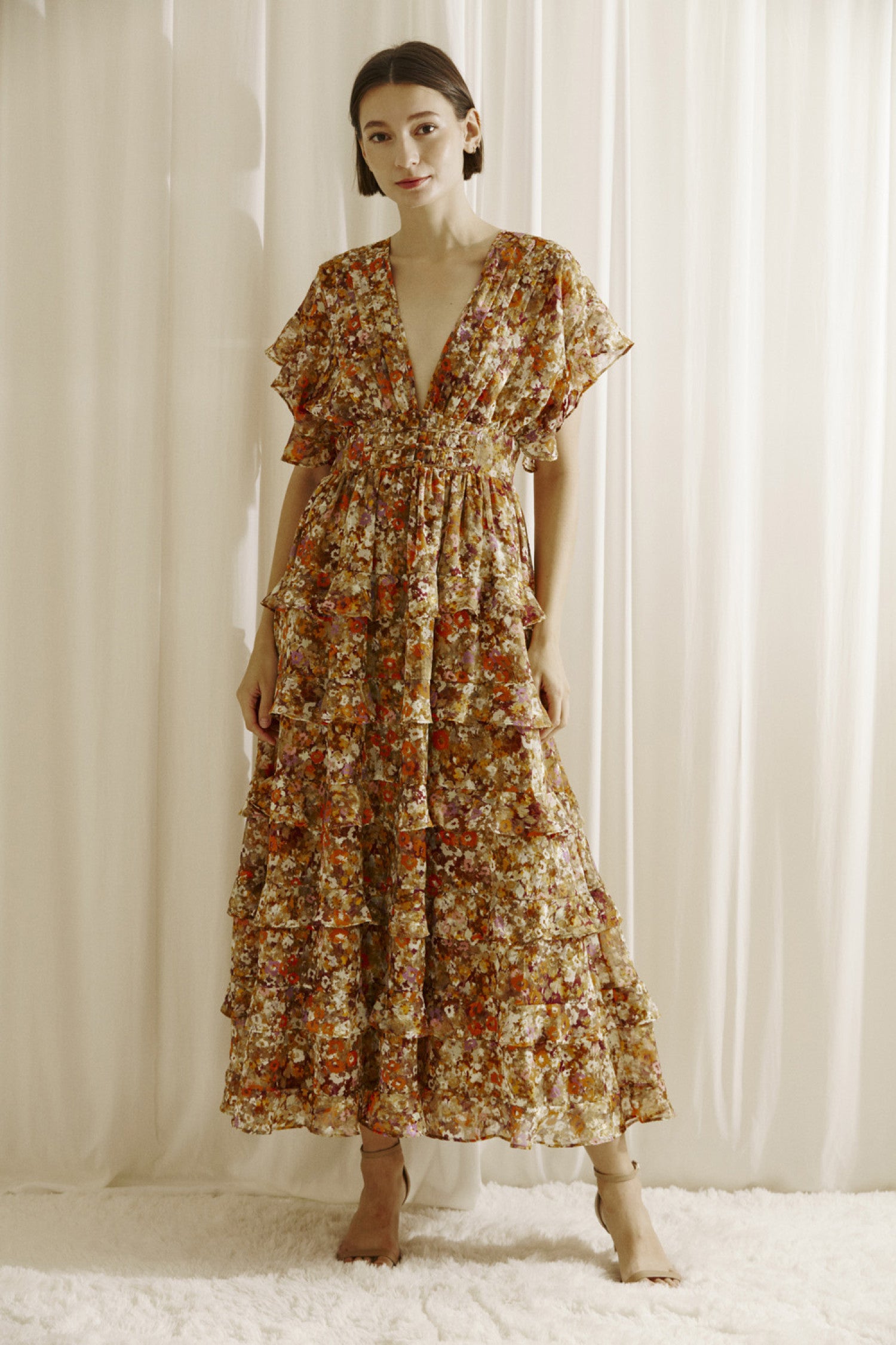 Ditsy floral print midi dress multicolor. Prominent browns, oranges, purples, and maroons.  It shows a deep v-neckline, short fluttered, ruffled sleeves, and a cinched empire waist with center buttons. It also has a ruffled, tiered layered, midi bottom and an open back with a crisscross back tie..  Front full view.