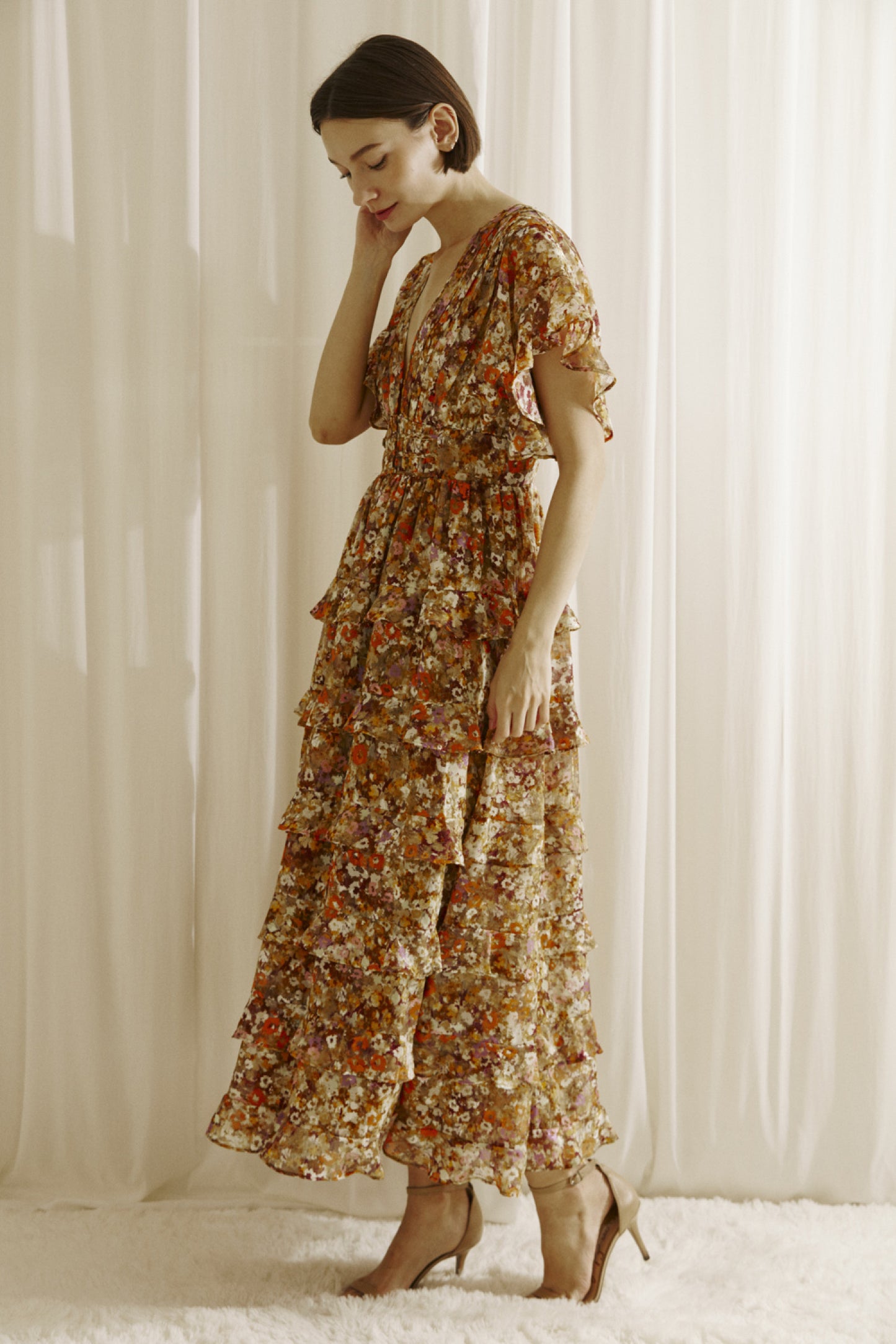 Ditsy floral print midi dress multicolor. Prominent browns, oranges, purples, and maroons.  It shows a deep v-neckline, short fluttered, ruffled sleeves, and a cinched empire waist with center buttons. It also has a ruffled, tiered layered, midi bottom and an open back with a crisscross back tie..  Sidel view.