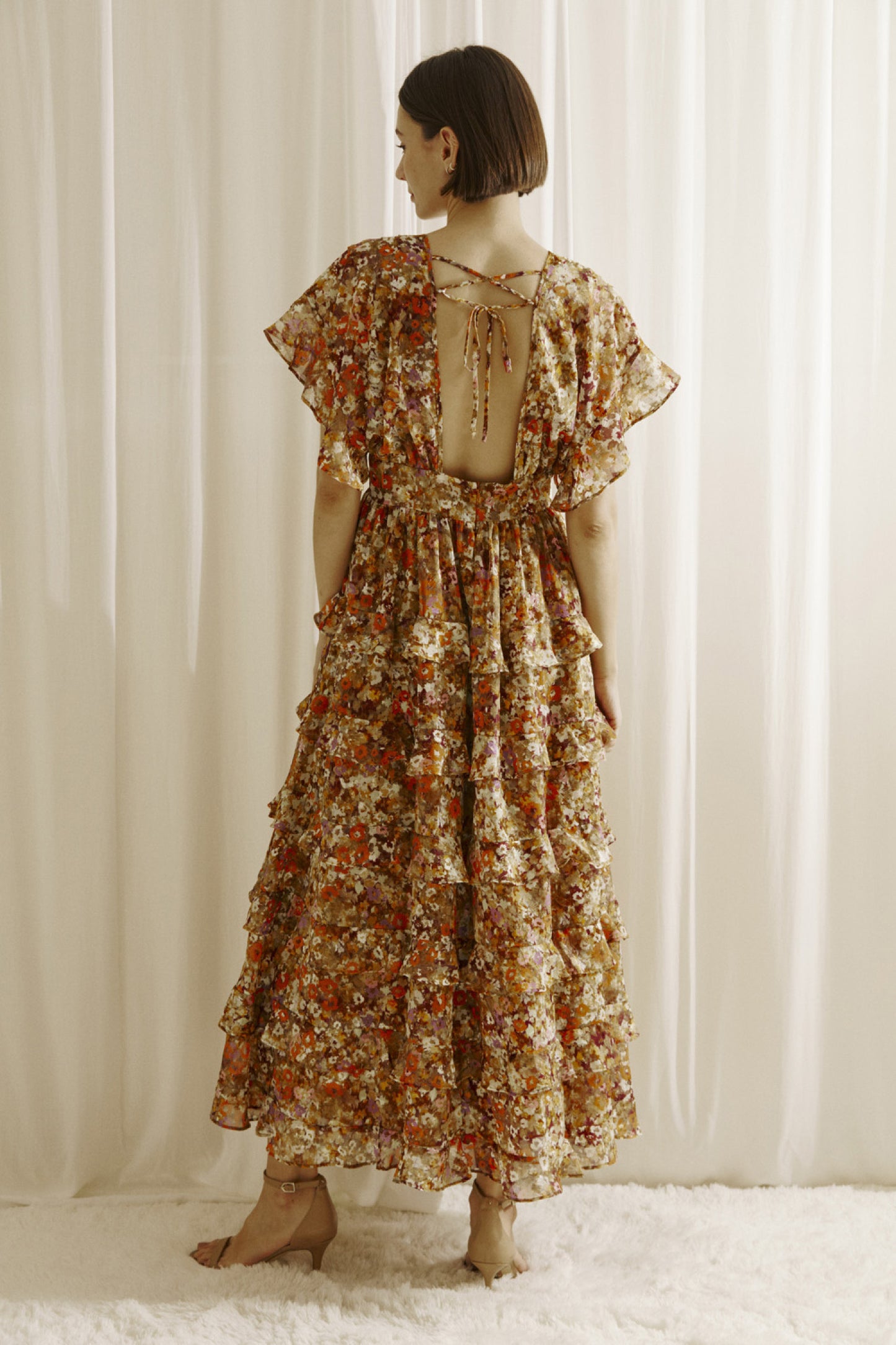 Ditsy floral print midi dress multicolor. Prominent browns, oranges, purples, and maroons.  It shows a deep v-neckline, short fluttered, ruffled sleeves, and a cinched empire waist with center buttons. It also has a ruffled, tiered layered, midi bottom and an open back with a crisscross back tie..  Back full view.
