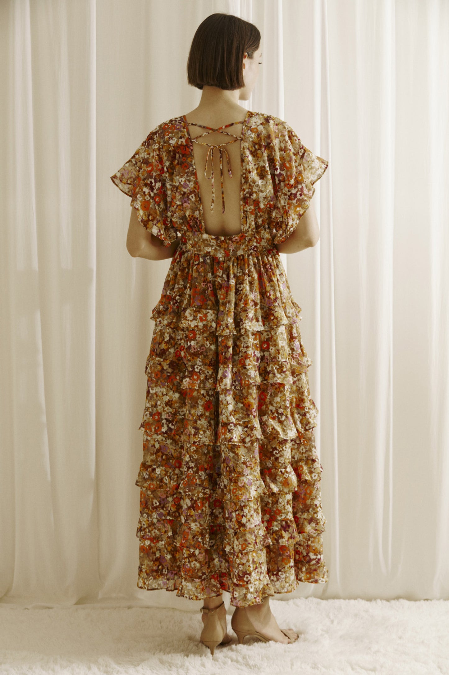 Ditsy floral print midi dress multicolor. Prominent browns, oranges, purples, and maroons.  It shows a deep v-neckline, short fluttered, ruffled sleeves, and a cinched empire waist with center buttons. It also has a ruffled, tiered layered, midi bottom and an open back with a crisscross back tie..  Back full view.